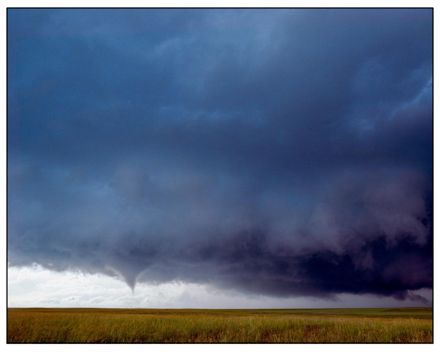 A tornado forms under a dark, stormy sky above a wide, grassy field.