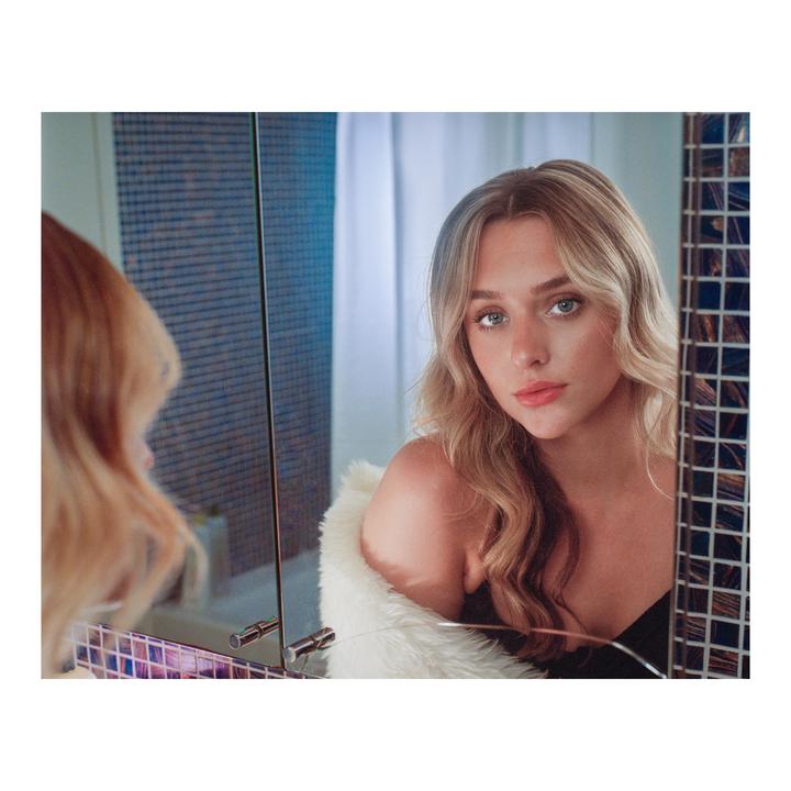 A woman with long wavy hair and a white fur stole looks at her reflection in a mirror.