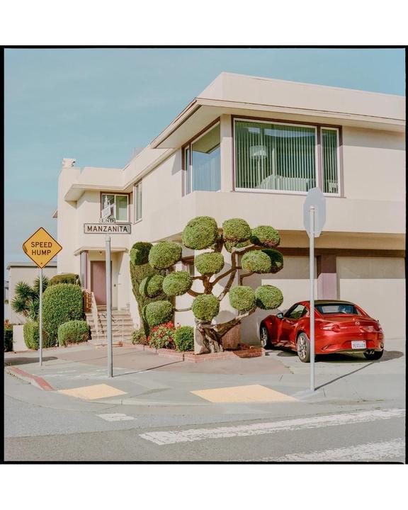 A red car is parked in front of a house with a manicured tree and a "Manzanita" street sign.