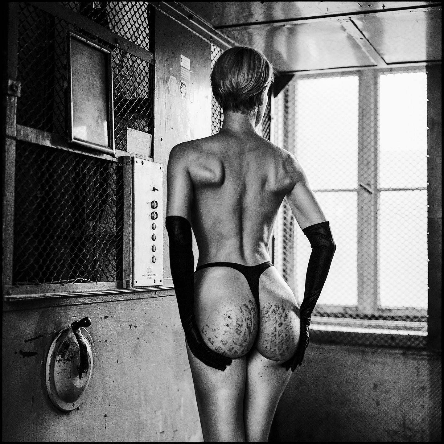 A woman in a thong and long gloves stands with her back to the camera in an elevator.