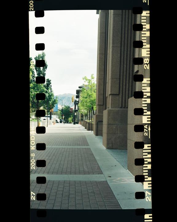 A wide sidewalk with brick pavers and a concrete border runs alongside a building with large columns and a street with trees and mountains in the distance.