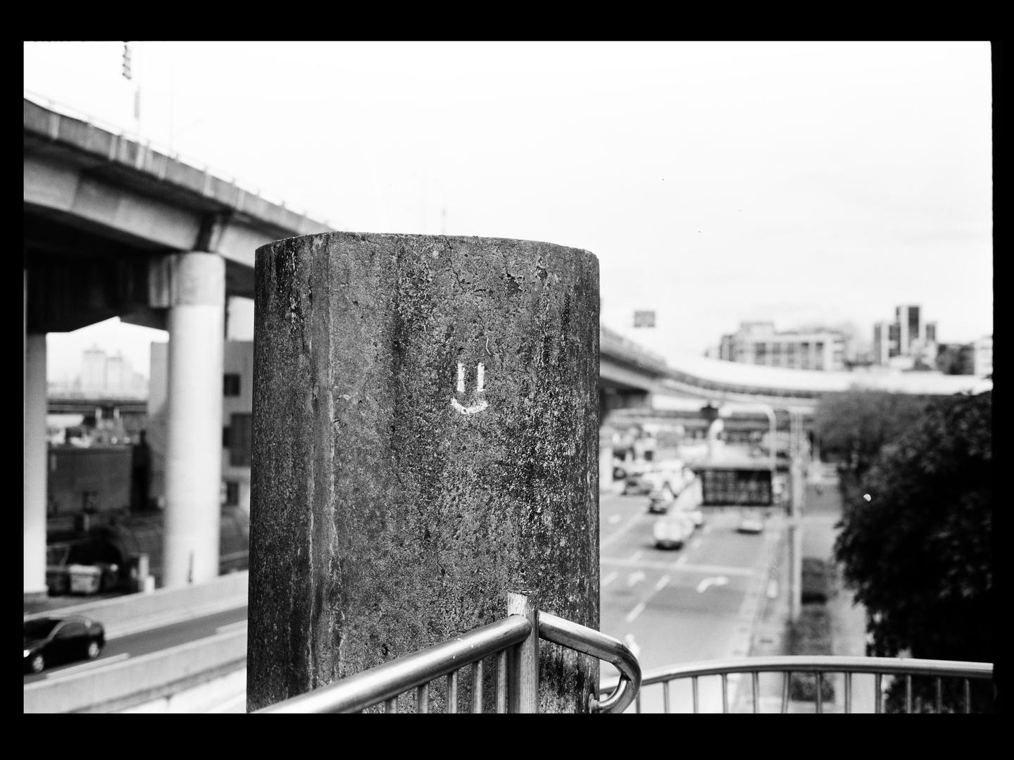 A smiley face is drawn on a concrete pillar next to a railing overlooking a city street with cars and an overpass.