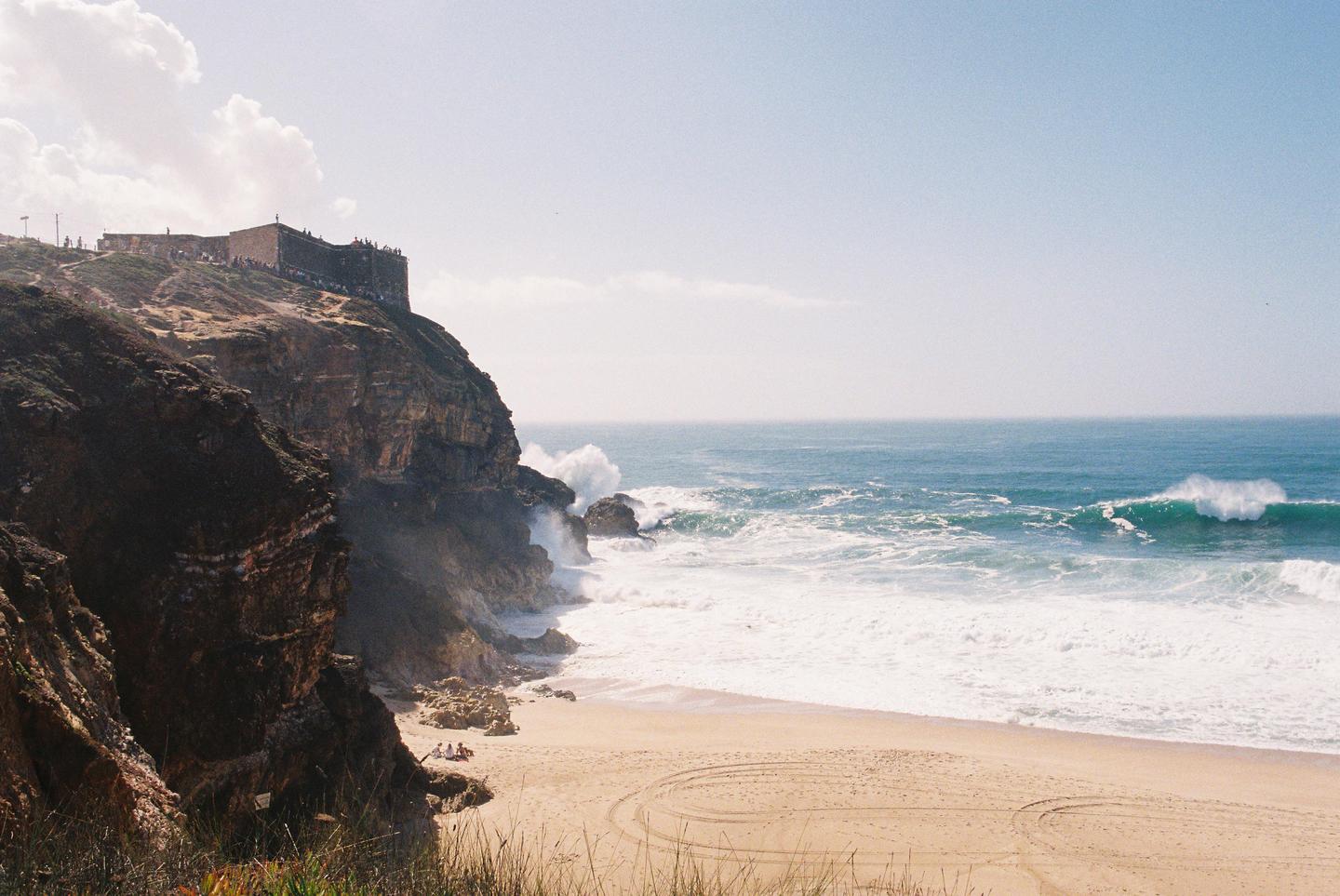 A large cliff with a building on top overlooks a sandy beach with crashing waves.