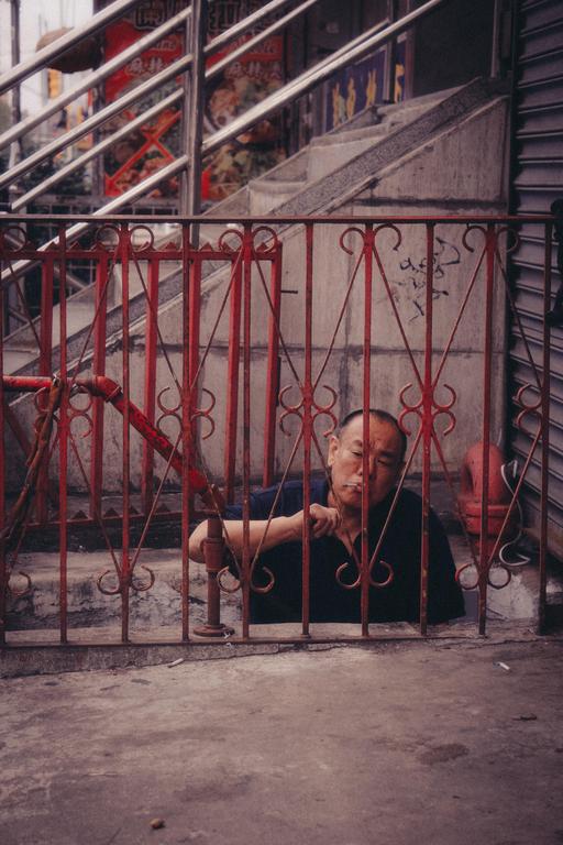 A man is visible behind a red metal fence on a street in Chinatown, New York City.