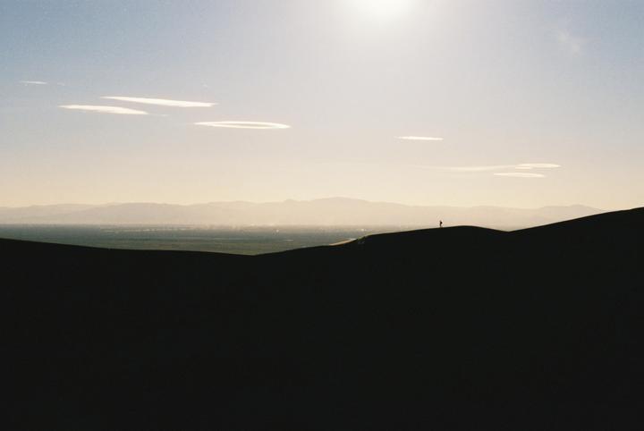 A person stands atop a sand dune with mountains and a valley in the distance under a bright sky.