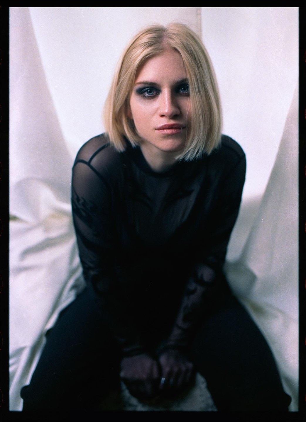 A woman with blonde hair and dark eye makeup sits looking directly at the viewer.
