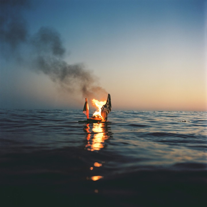 A small burning boat floats on the water at dusk, leaving a trail of smoke.