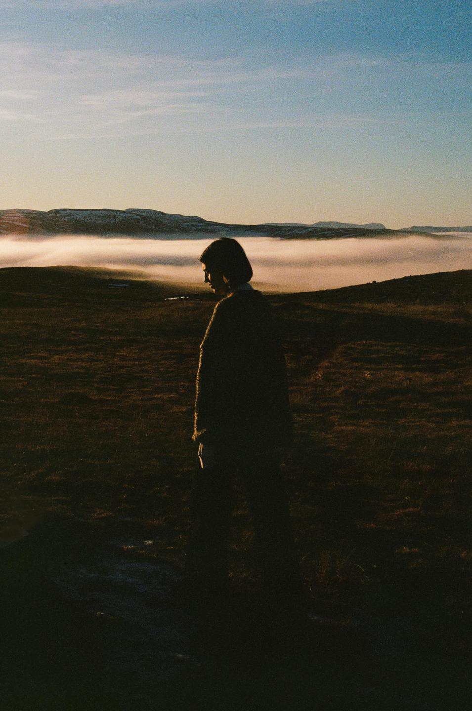 A woman stands in a field with a layer of fog in the distance and mountains under a clear sky.