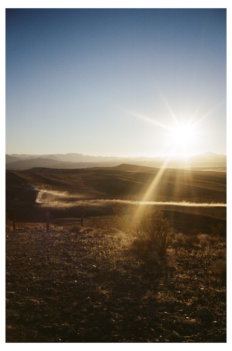 The sun shines brightly over a desert landscape with mountains in the distance and a dust cloud on the horizon.