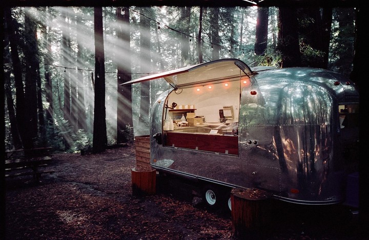 A shiny silver food truck with its serving window open is parked in a forest with sun rays shining through the trees.