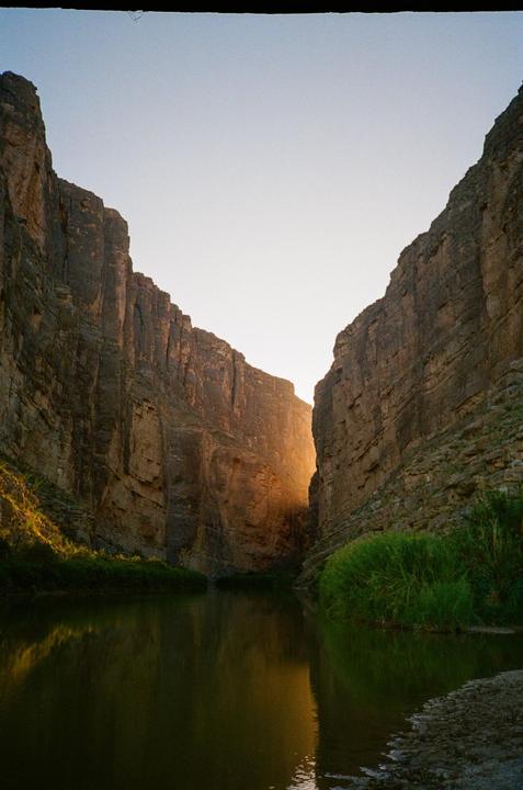A river flows through a canyon with tall rock walls and green vegetation on its banks, illuminated by a bright light source in the distance.