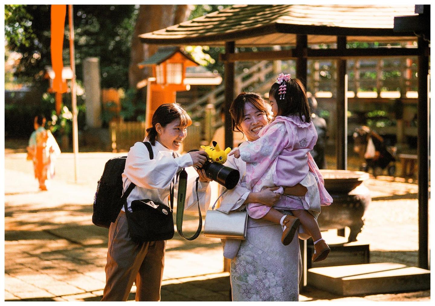 Two women and a child are smiling and looking at each other in a sunlit outdoor setting with traditional Japanese architecture.