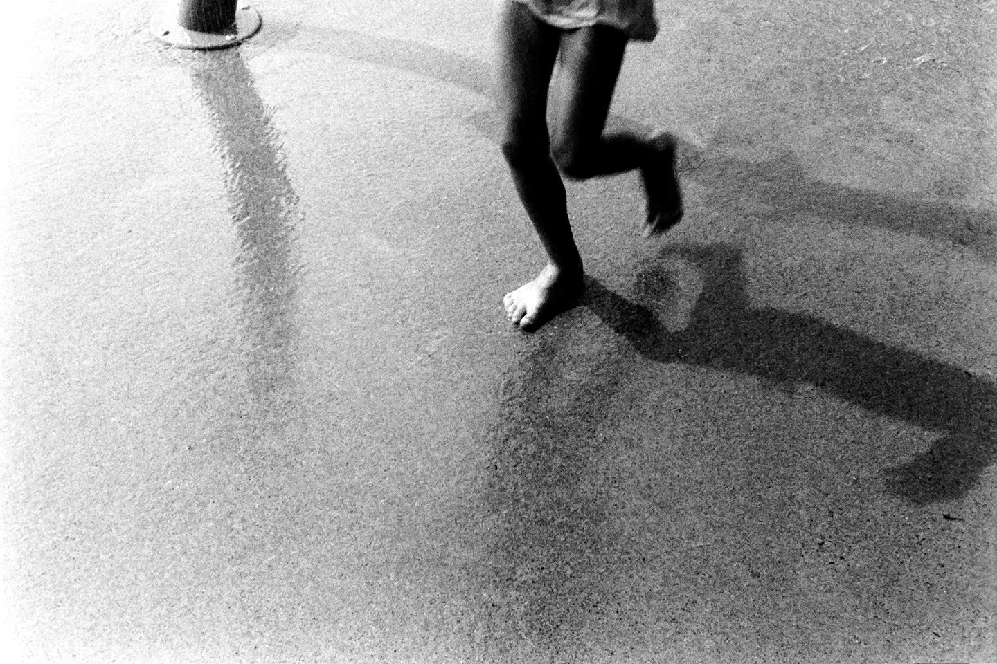 A child's legs and feet are visible as they run across a wet, textured surface, casting a long shadow.