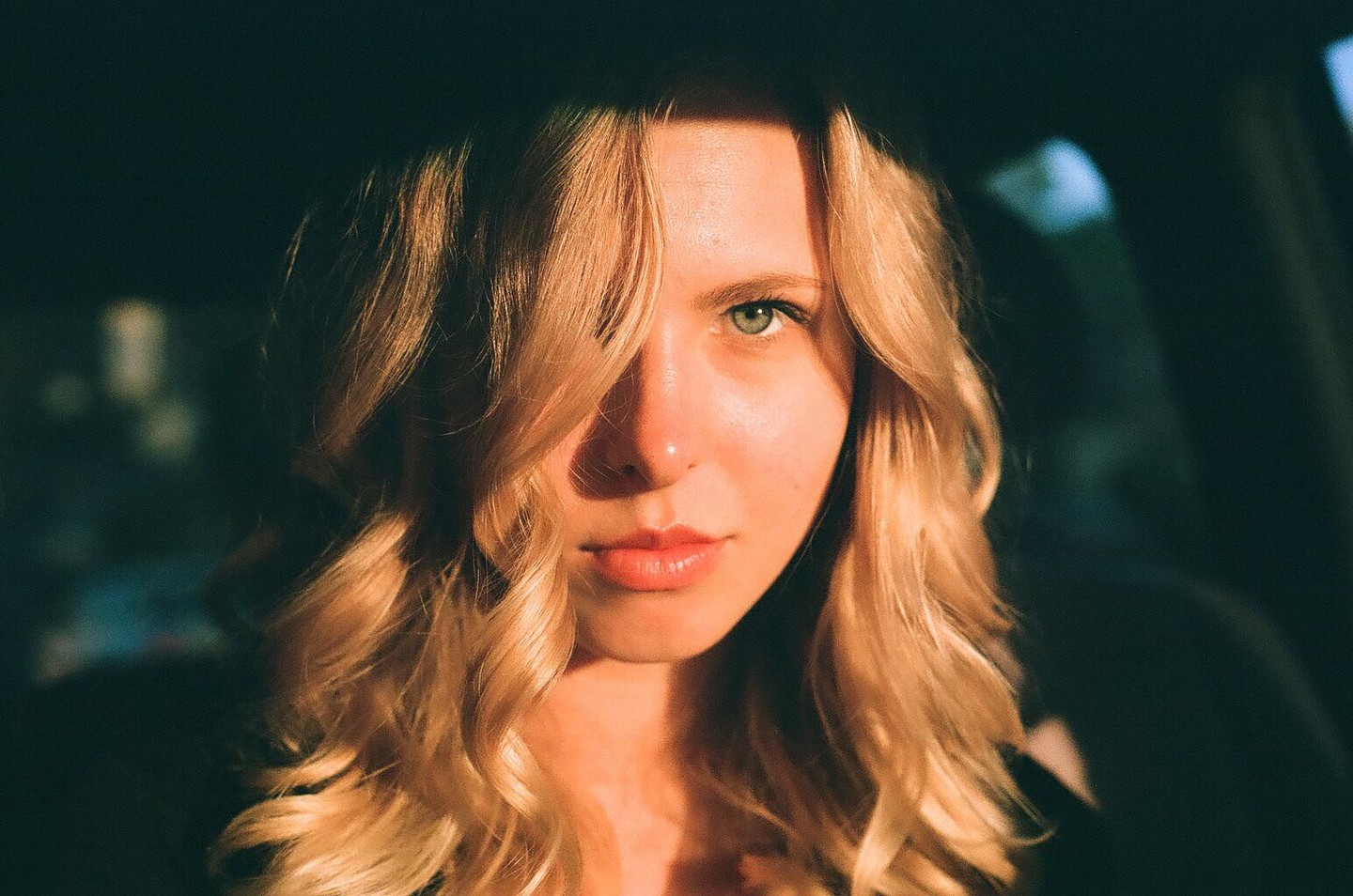 A woman with blonde hair and green eyes looks directly at the viewer, with part of her face in shadow.