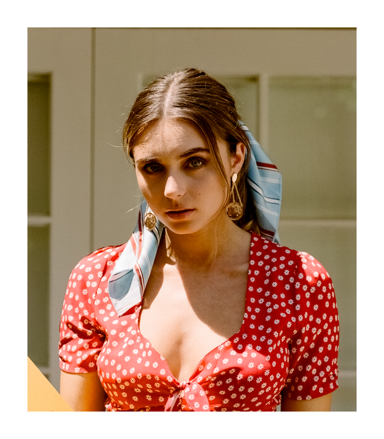 A woman wearing a red floral top, a headscarf, and coin earrings looks directly at the viewer.