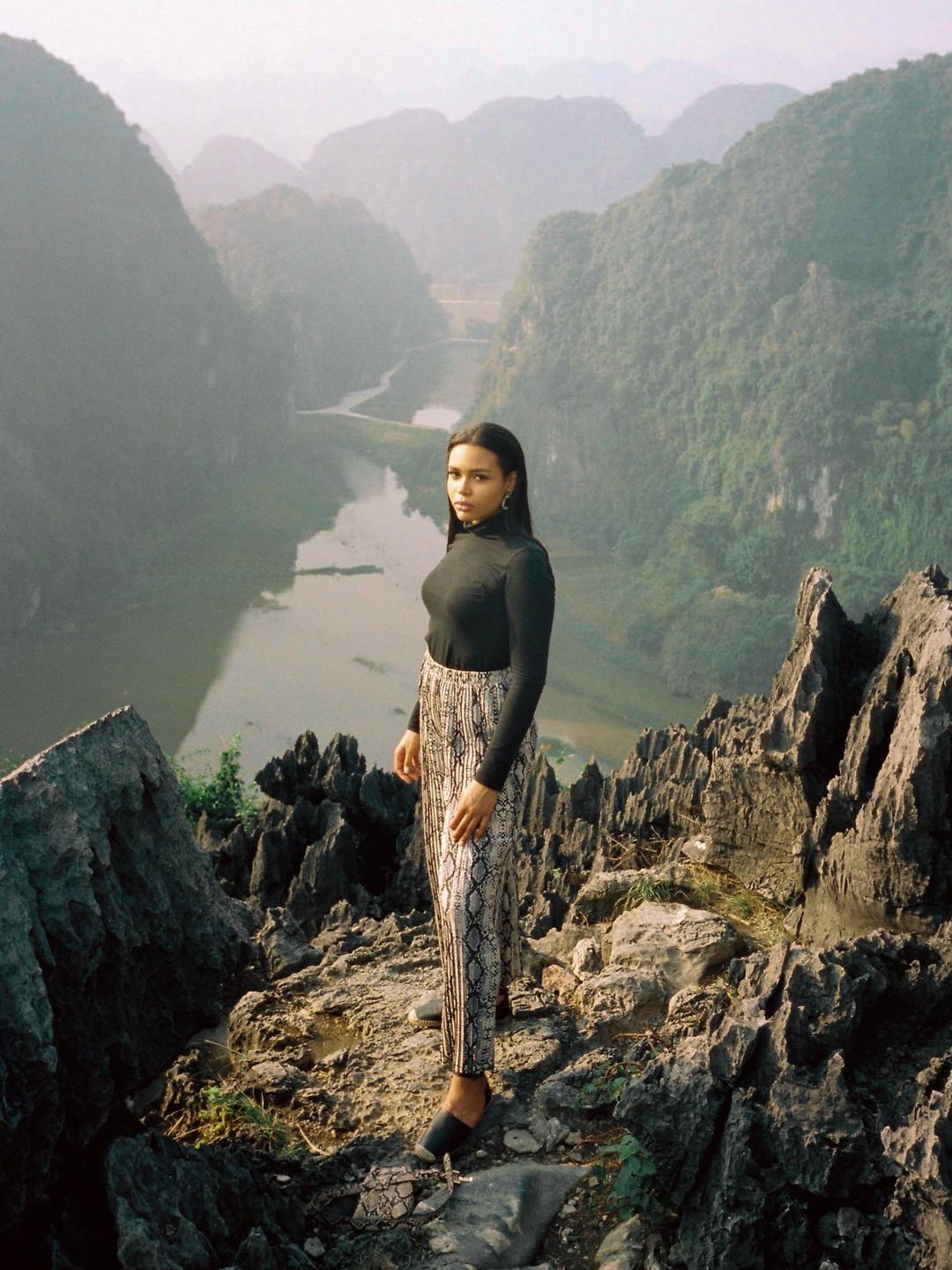 A woman stands on a rocky mountain overlooking a river winding through a valley.