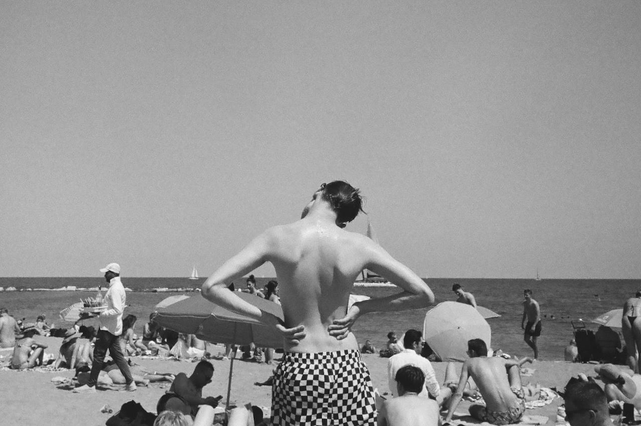 A person with their back to the camera stands on a crowded beach looking out at the ocean.