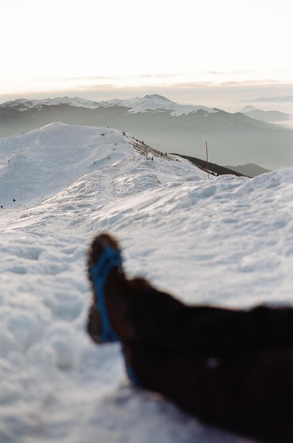 A person's legs and feet are visible in the foreground of a snowy mountain landscape.