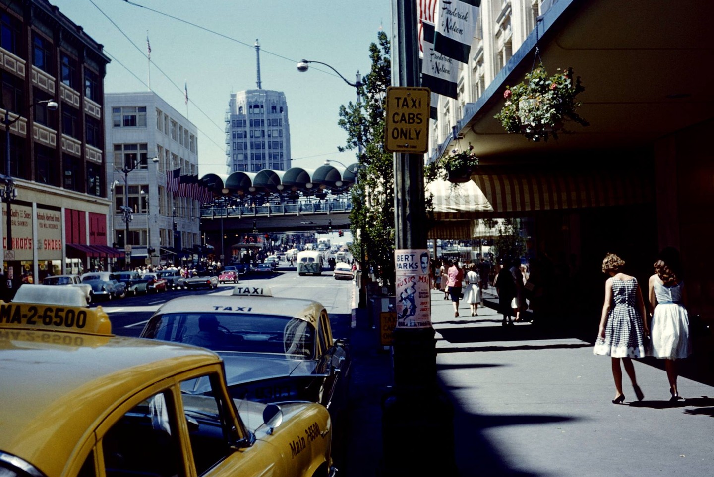 Two women walk along a sunlit sidewalk past parked taxis and a busy street with a tall building in the distance.