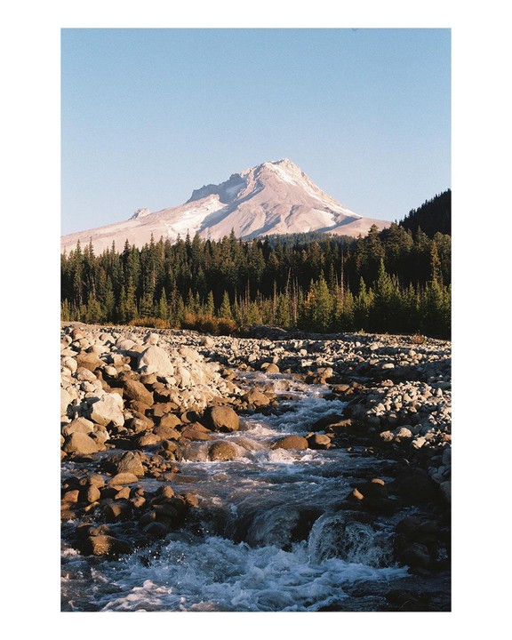 A river flows over rocks in the foreground, with a forest and a snow-capped mountain in the background.
