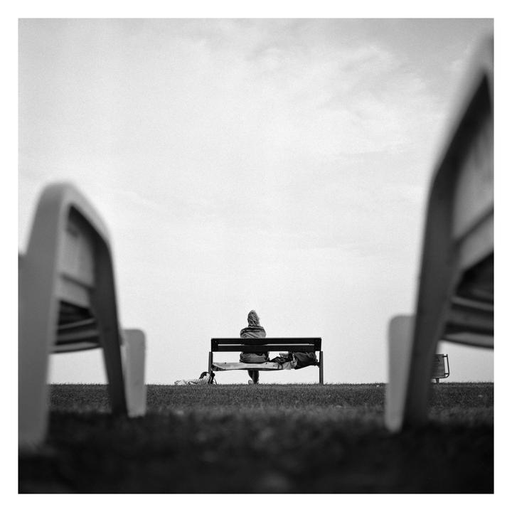 A person sits alone on a bench in a grassy field, viewed from a low angle between two other benches.