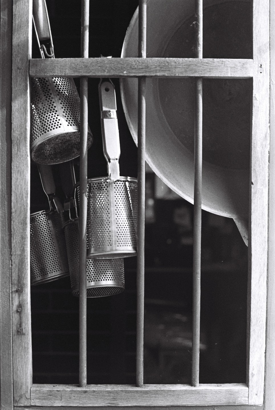 Several metal noodle strainers hang behind a wooden window frame with vertical bars.