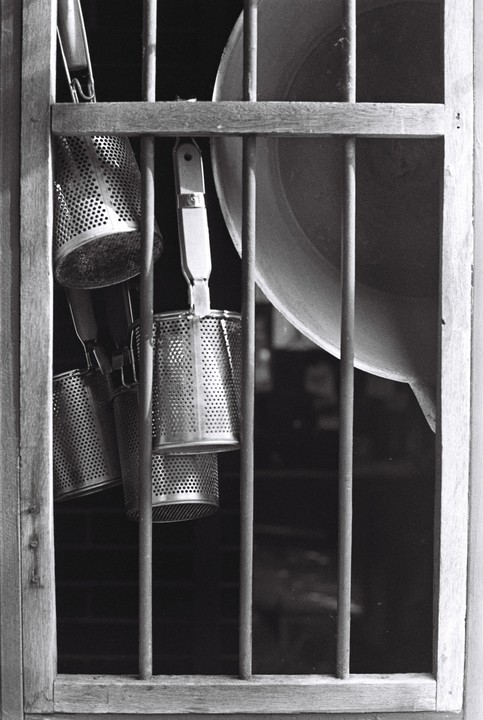 Several metal noodle strainers hang behind a wooden window frame with vertical bars.