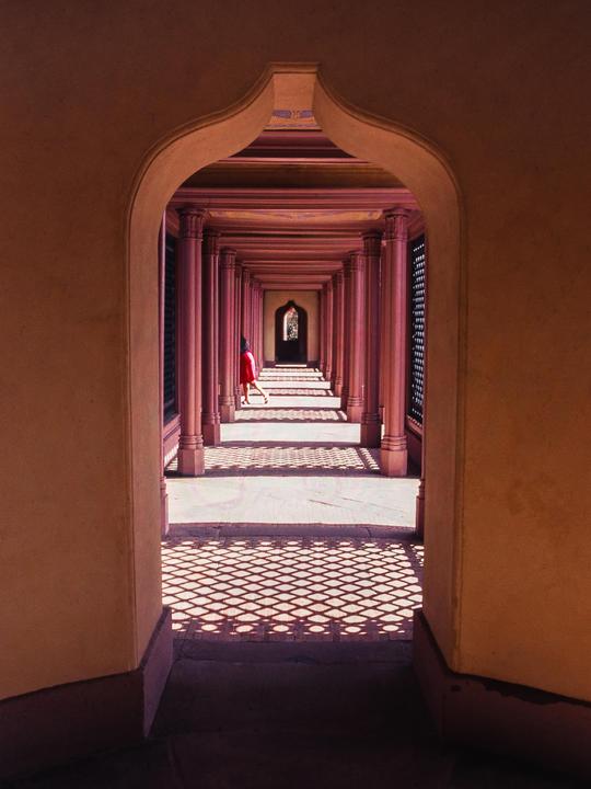 A person in a red dress walks down a long hallway with columns and patterned shadows on the floor.