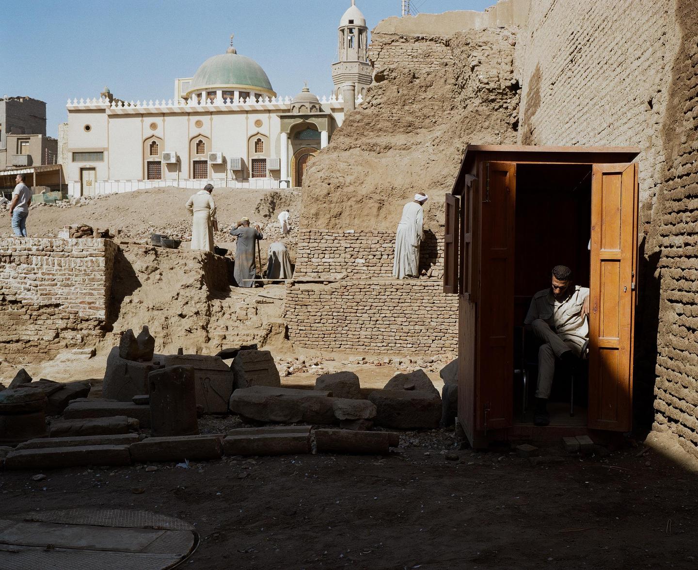 A man sits in a wooden booth while other men work at an excavation site with a mosque in the background.