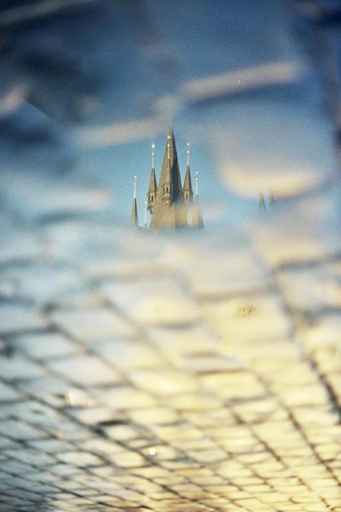 A castle with multiple spires is reflected in a puddle on a cobblestone street.