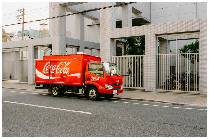 A red Coca-Cola truck is parked on the side of a street next to a modern building with a tiled facade.