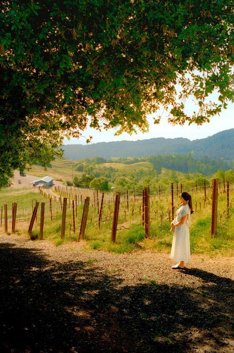 A woman stands on a gravel path next to a vineyard with rolling hills and a barn in the background.