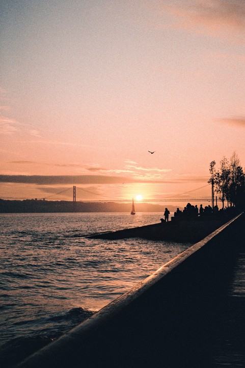 A sailboat passes in front of a bridge as the sun sets over a body of water with people gathered on the shore.