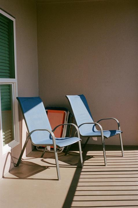Two blue chairs sit on a patio with strong shadows from a nearby window.