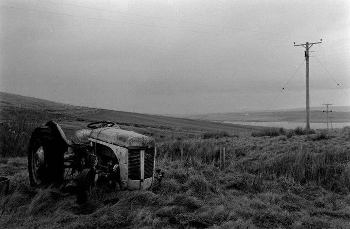 An old tractor sits in a field of tall grass with a body of water and hills in the background under a cloudy sky.