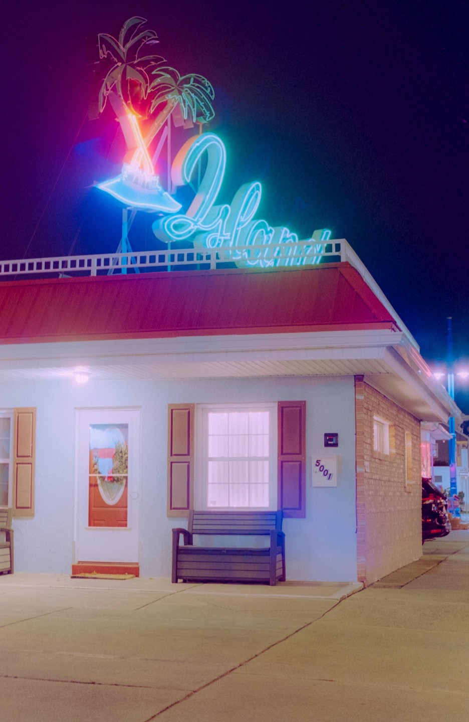 A neon sign with palm trees and a cocktail glass sits atop a building with a red roof and a bench in front of a window.