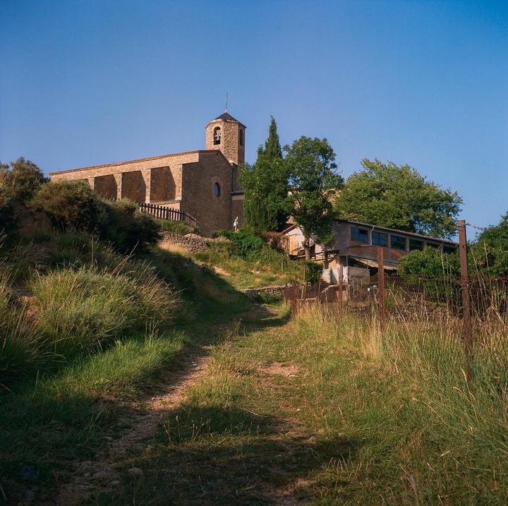 A dirt path leads up a grassy hill to a stone church with a bell tower and a building with a fence.