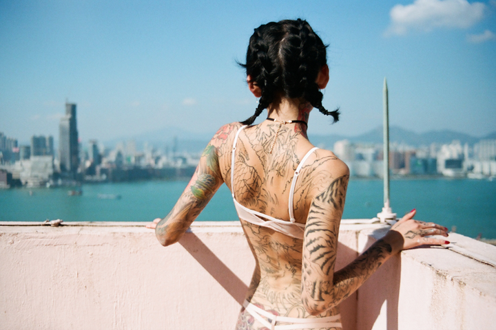 A woman with braided hair and extensive tattoos on her back and arms stands on a balcony overlooking a city and body of water.