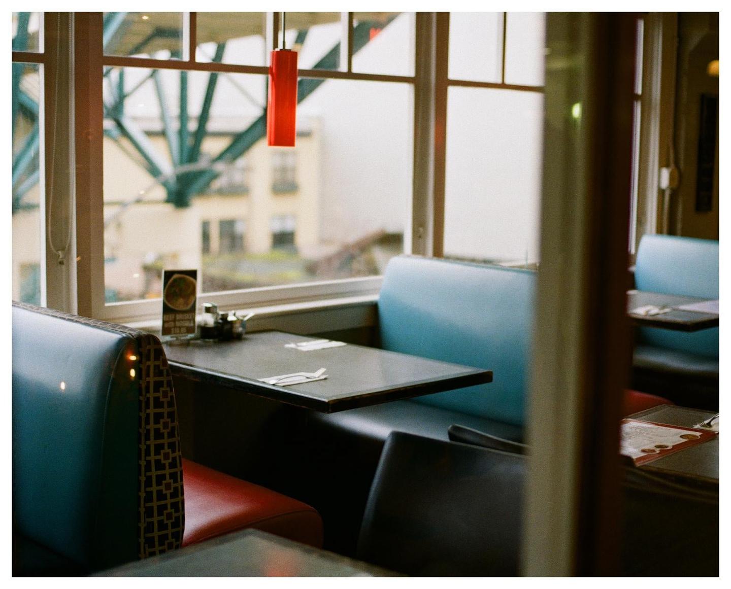 A diner booth with blue and red upholstery is visible next to a window looking out onto a city.
