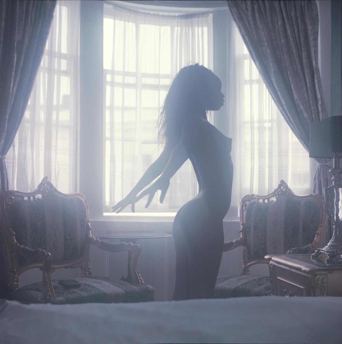 A nude woman stands in profile with her arms outstretched in a room with two ornate chairs and a window.
