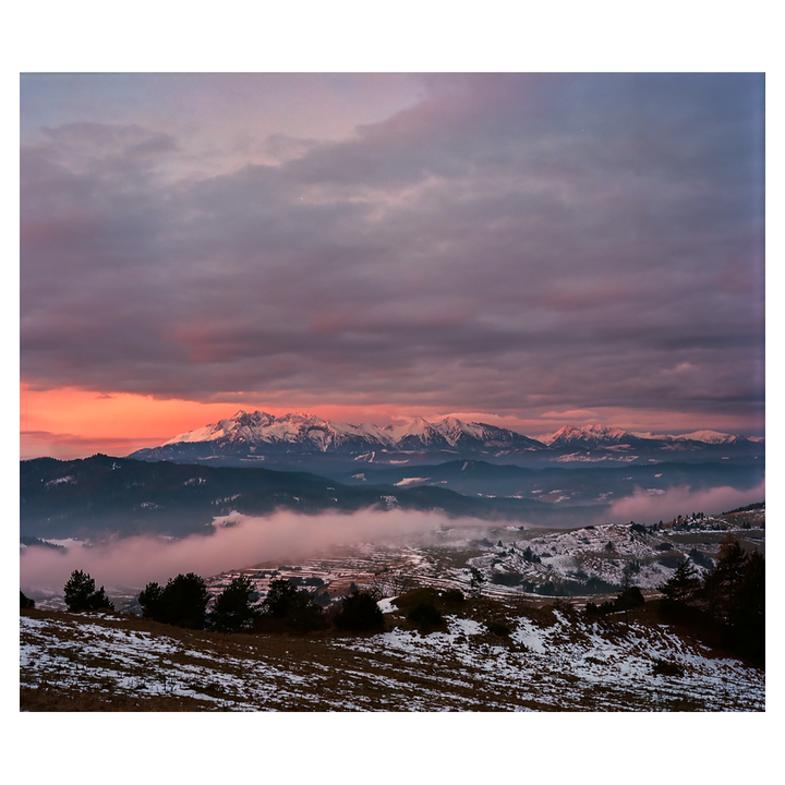 A mountain range with snow-capped peaks is visible under a dramatic sky with pink and purple clouds, with fog covering the valley below.