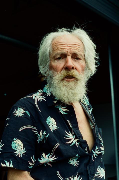 A man with a long white beard and white hair wears an open Hawaiian shirt with pineapple and palm tree patterns.