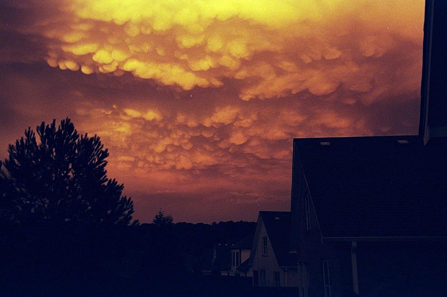 A dramatic sky with mammatus clouds hangs over silhouetted houses and trees at dusk.