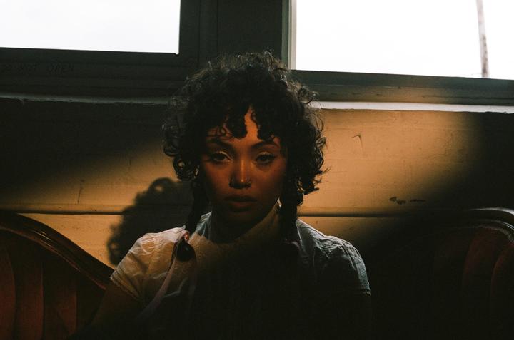 A woman with curly hair and braids sits in a dimly lit room, looking directly at the viewer.
