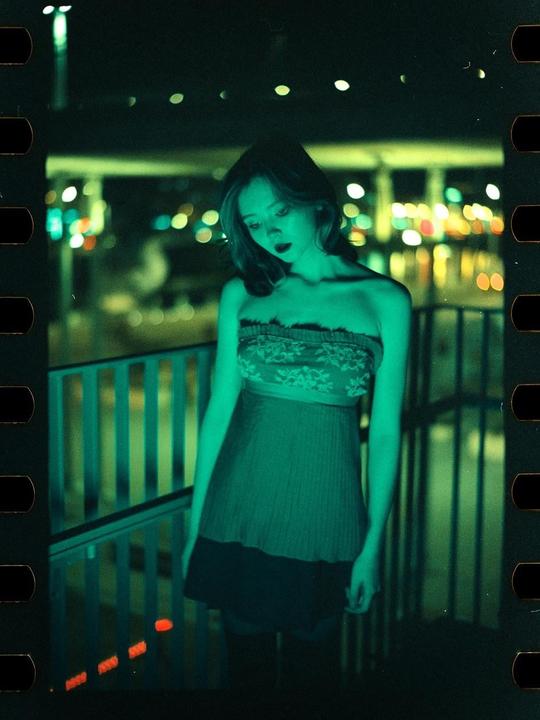A woman in a strapless dress stands on a balcony at night, illuminated by green light.