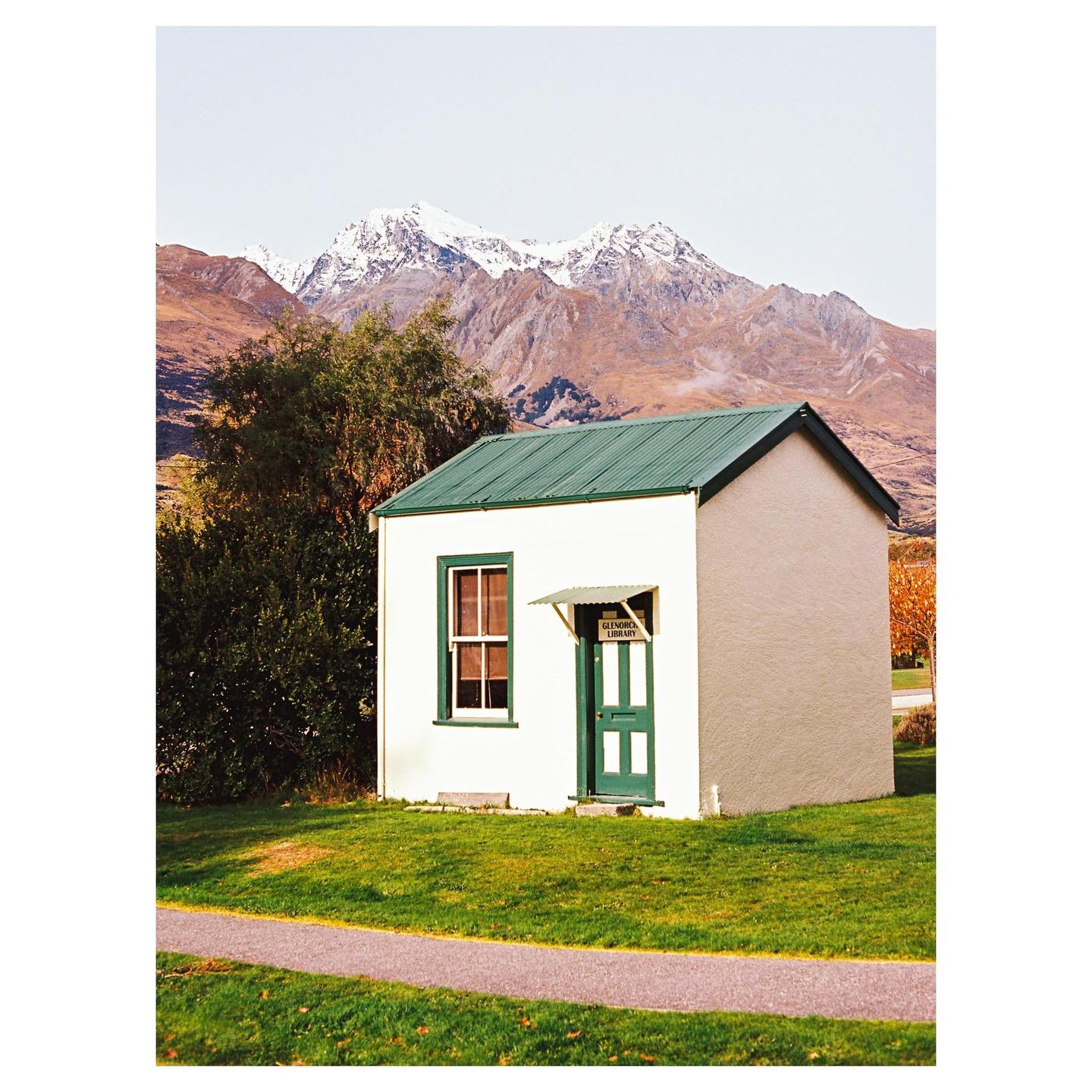 A small white building with a green roof and door stands in a grassy field with snow-capped mountains in the background.