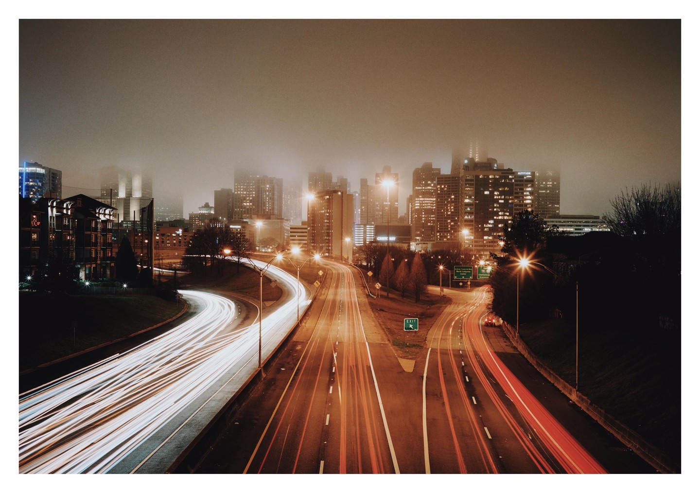 A highway with light trails from vehicles leads into a hazy city skyline at night.