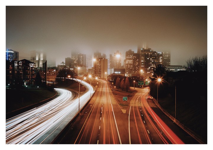 A highway with light trails from vehicles leads into a hazy city skyline at night.