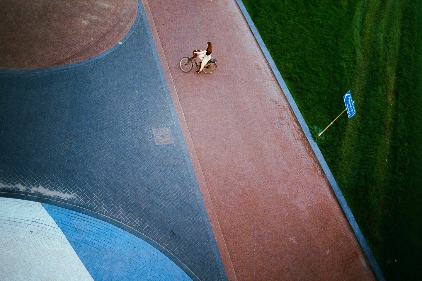 A woman rides a bicycle on a paved path next to a grassy area with a blue sign.