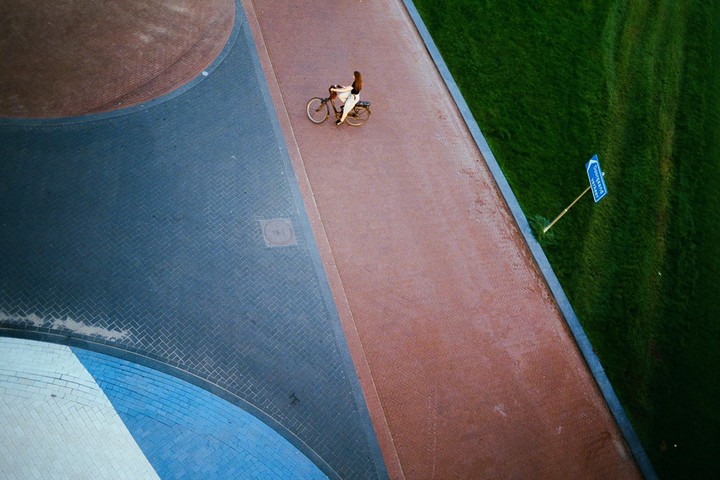 A woman rides a bicycle on a paved path next to a grassy area with a blue sign.